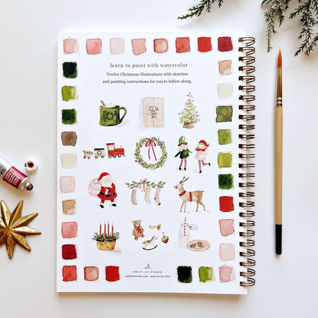 Spiral-bound art book with Christmas illustrations on a white surface with a paintbrush and watercolor bottles.