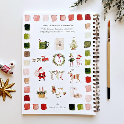 Spiral-bound art book with Christmas illustrations on a white surface with a paintbrush and watercolor bottles.