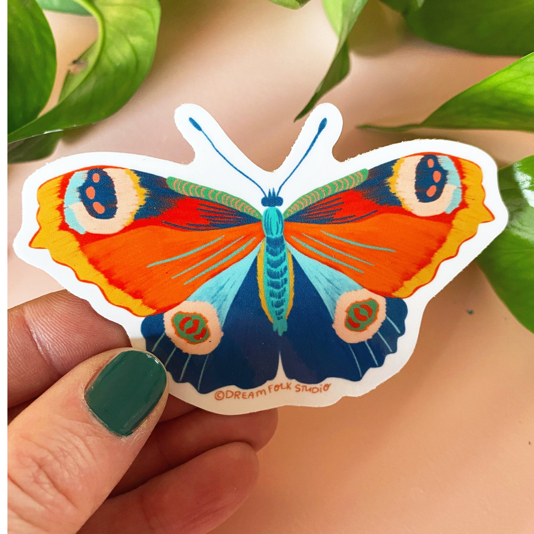 Colorful butterfly sticker held by a hand with green nail polish against a natural background.