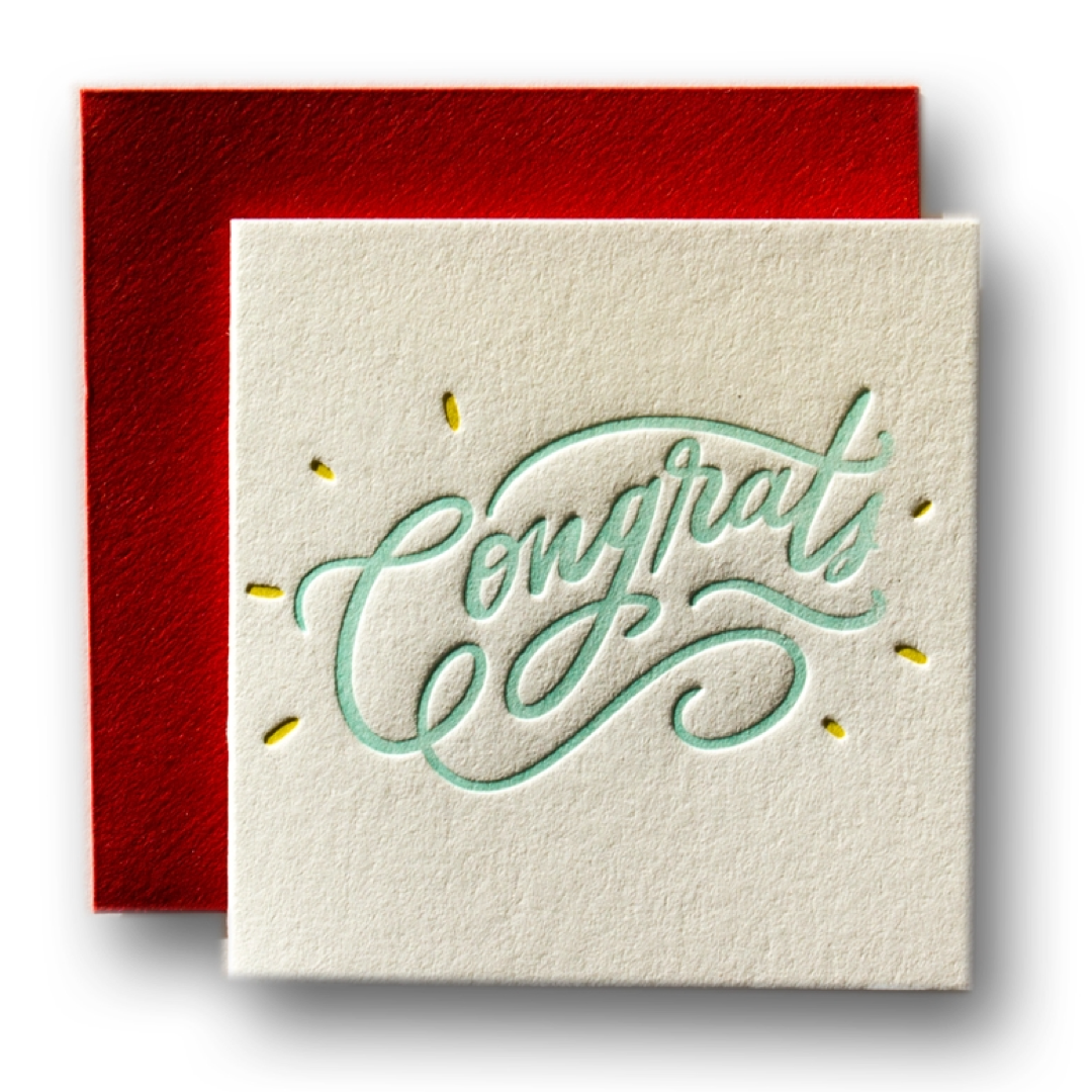 A square congratulations card in textured off-white paper with the word 'Congrats' in light mint green cursive and small gold dot accents, shown with a red envelope behind it on a white background.
