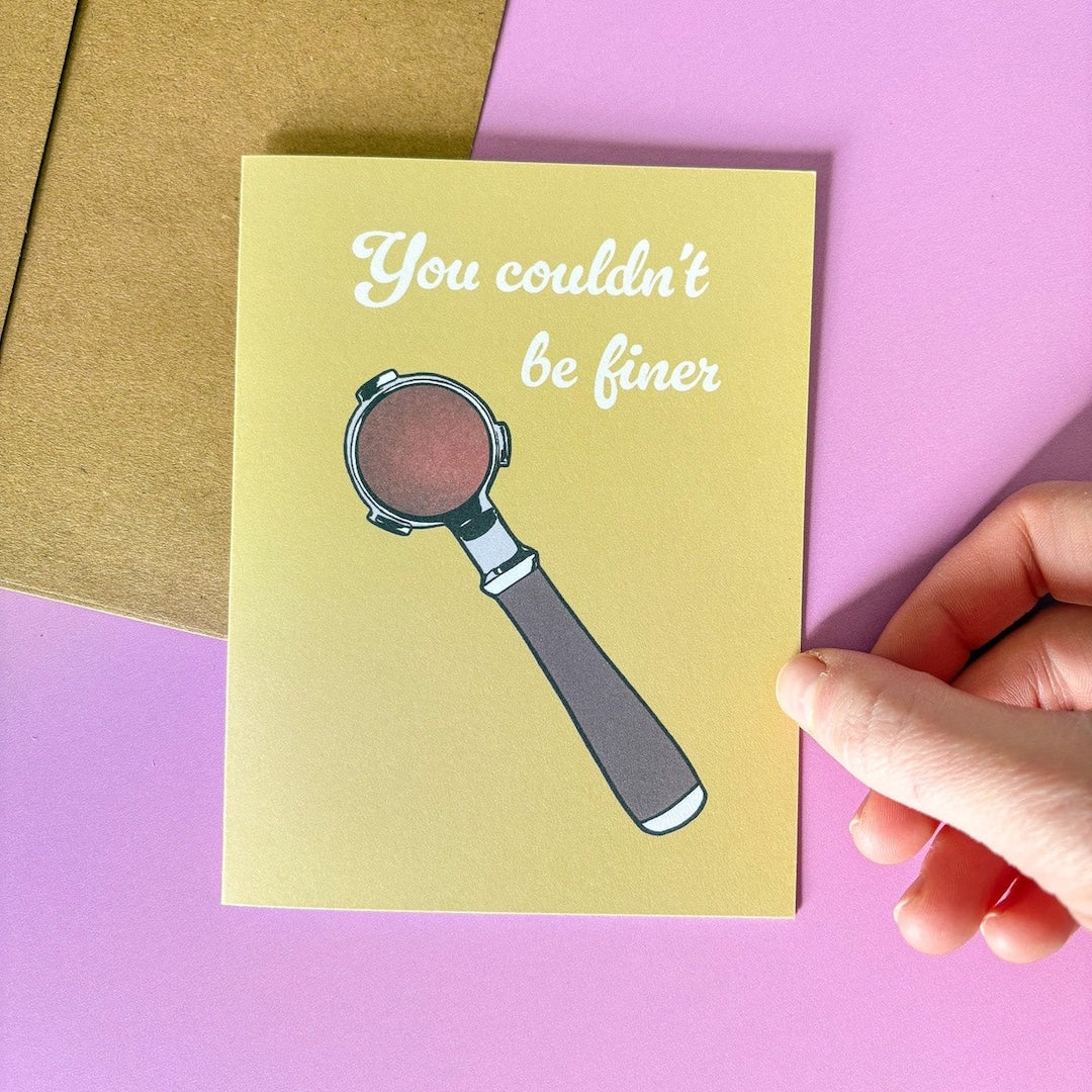 Greeting card with an espresso portafilter illustration and text that says 'you couldn't be finer,' held by a hand on a pink and gold background.