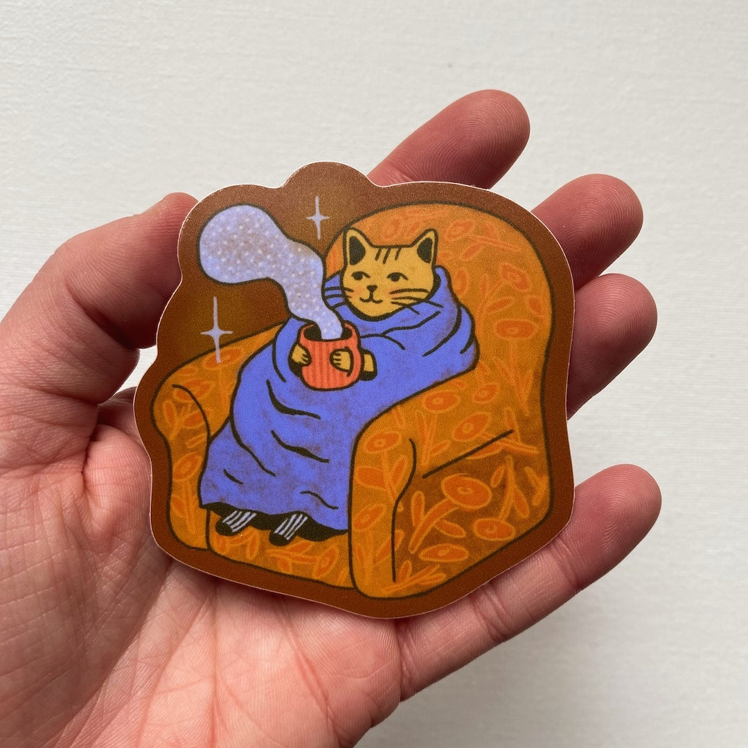 Hand holding a sticker of a cat sitting on a chair with a mug, wrapped in a blanket