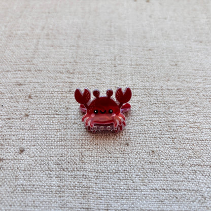 A small red crab-shaped hair clip displayed on a textured beige fabric background.