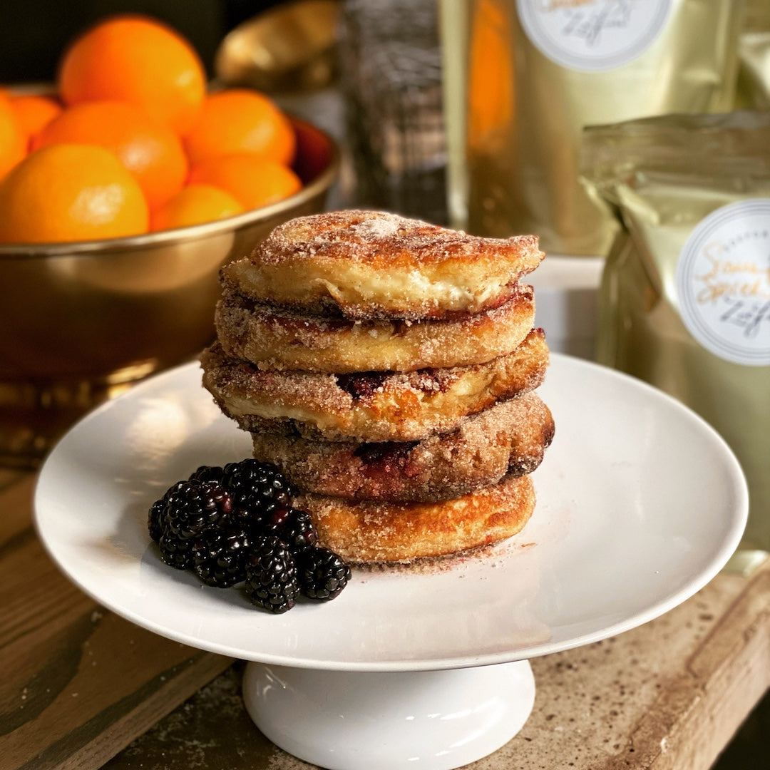 A stack of four cinnamon sugar pancakes  sits on a white pedestal plate, garnished with blackberries, with oranges and packaged goods blurred in the background.