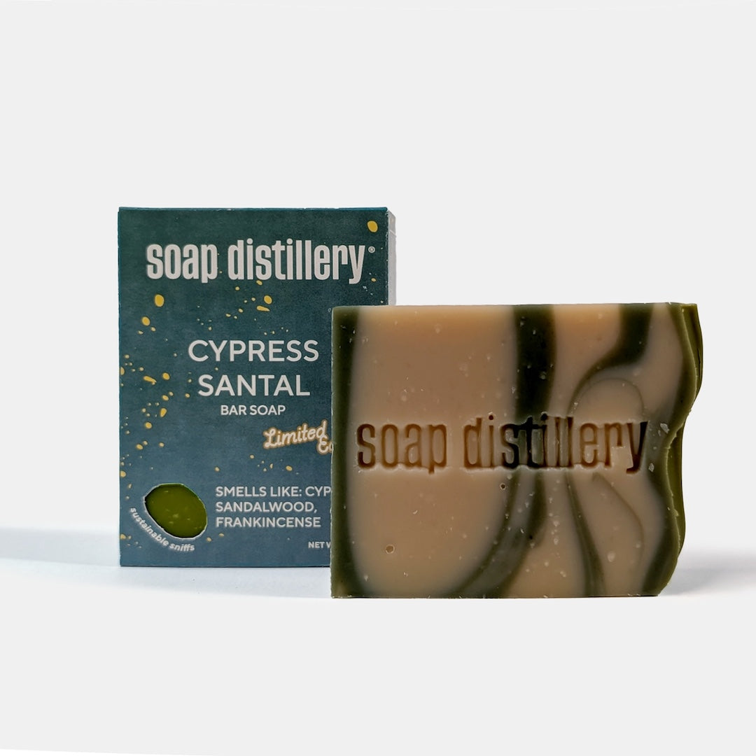A bar of soap and its packaging box from the brand 'soap distillery'. The box is dark teal with white text and gold speckles, labeled 'CYPRESS SANTAL BAR SOAP'.
