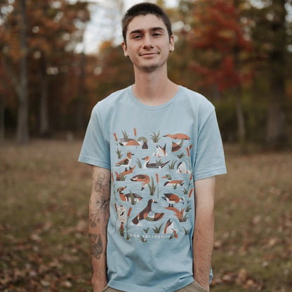 A man wearing a light blue graphic t-shirt with a detailed print of various ducks  standing in an outdoor park with autumn trees in the background.