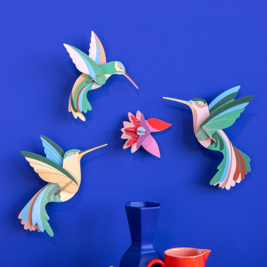 Three colorful 3D cardboard hummingbird wall sculptures and a matching flower ornament displayed on a vibrant blue wall, with a dark blue vase and a red cup partially visible at the bottom.