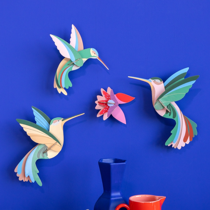 Three colorful 3D cardboard hummingbird wall sculptures and a matching flower ornament displayed on a vibrant blue wall, with a dark blue vase and a red cup partially visible at the bottom.