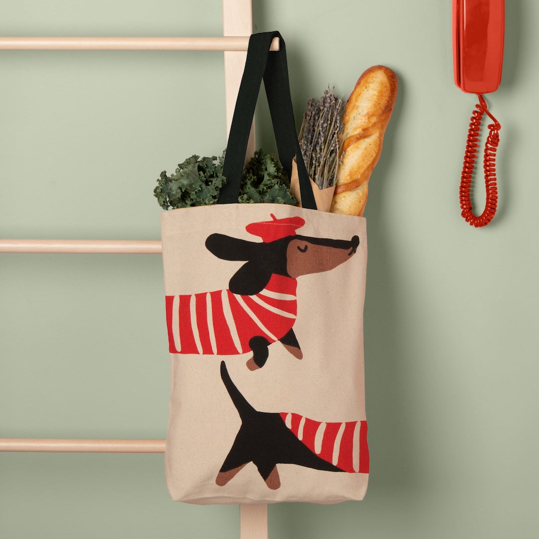 Tote bag with dachshund design hanging on a hook against a green wall.