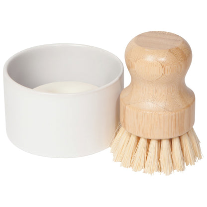 A natural bamboo-handled cleaning brush with light beige bristles next to a white ceramic cup containing a small amount of  soap, on a solid white background.
