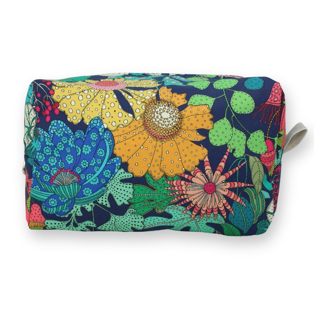 A rectangular cosmetic pouch with a colorful, busy floral and botanical pattern on a navy blue background, featuring a small white strap on the right side.