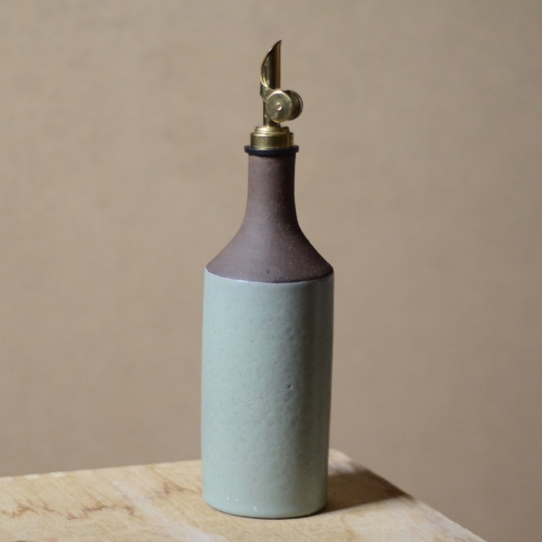 Two-toned brown and green ceramic oil bottle with brass nozzle on a beige surface.