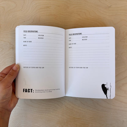 An open bird journal with printed observation pages held in a hand over a light wood surface. The pages contain sections for about the weather, name of bird, notes, location, time, and a space to sketch the bird