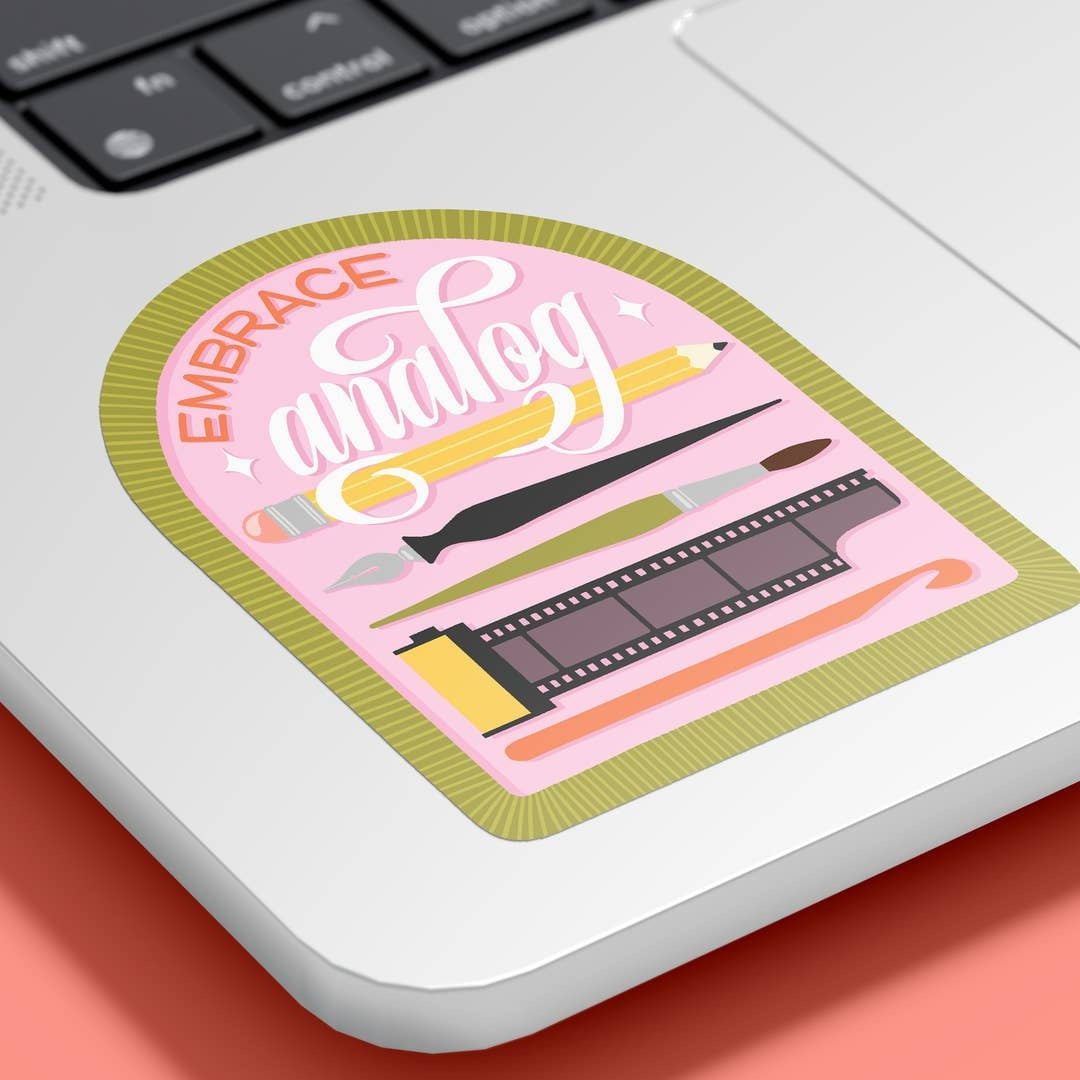 A die-cut sticker with the text 'EMBRACE analog' and illustrated art tools on a pink background, placed on a silver laptop palm rest over a coral surface.