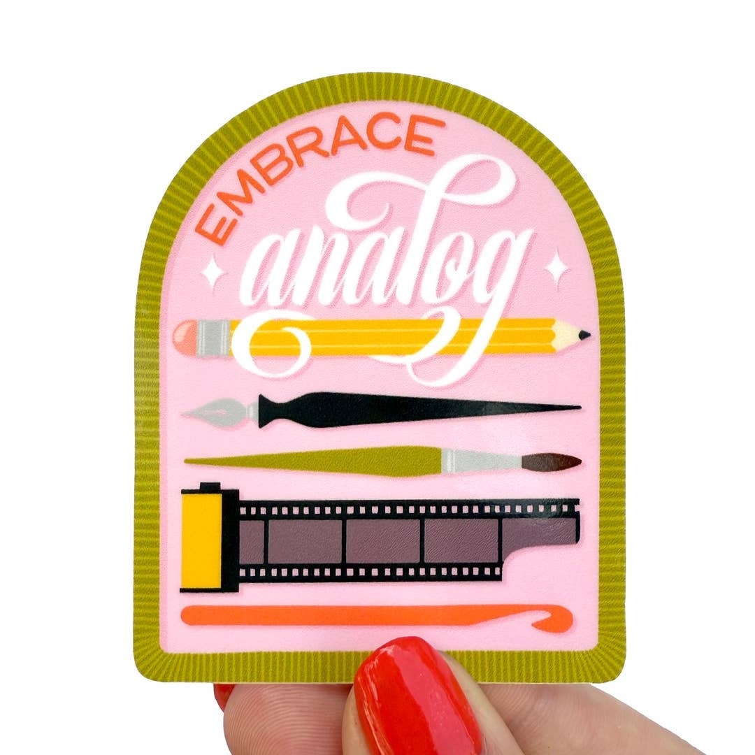 A hand with red painted nails holds a die-cut sticker featuring the words 'EMBRACE analog' in orange and white script on a pink background, surrounded by illustrated art tools like a pencil, paintbrush, and film strip. The sticker has an olive green border.