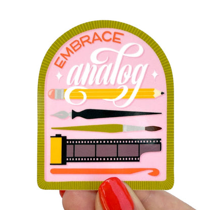 A hand with red painted nails holds a die-cut sticker featuring the words 'EMBRACE analog' in orange and white script on a pink background, surrounded by illustrated art tools like a pencil, paintbrush, and film strip. The sticker has an olive green border.
