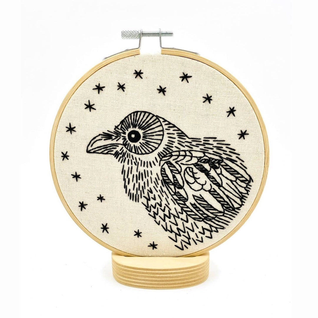 A circular embroidery hoop with a black line-art illustration of a raven surrounded by small black stars. The hoop is mounted on a matching wooden stand against a plain white background.