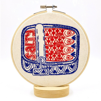 A circular embroidery hoop mounted on a wooden stand, featuring  design of a sardine can with red fish, blue borders, and the word 'SALT' in red on an off-white fabric background.
