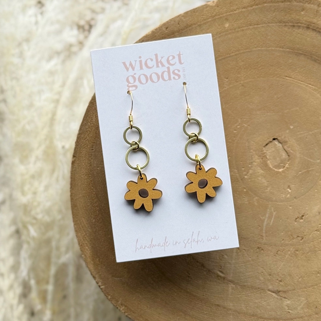 Gold flower-shaped earrings on a 'Wicket Goods' card against a wooden background