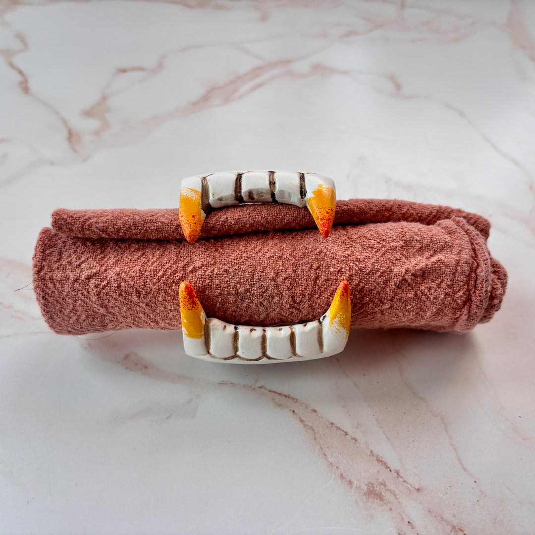 A rolled terracotta-colored textured towel or napkin secured with a vampire fang shaped napkin ring on a white marble surface.