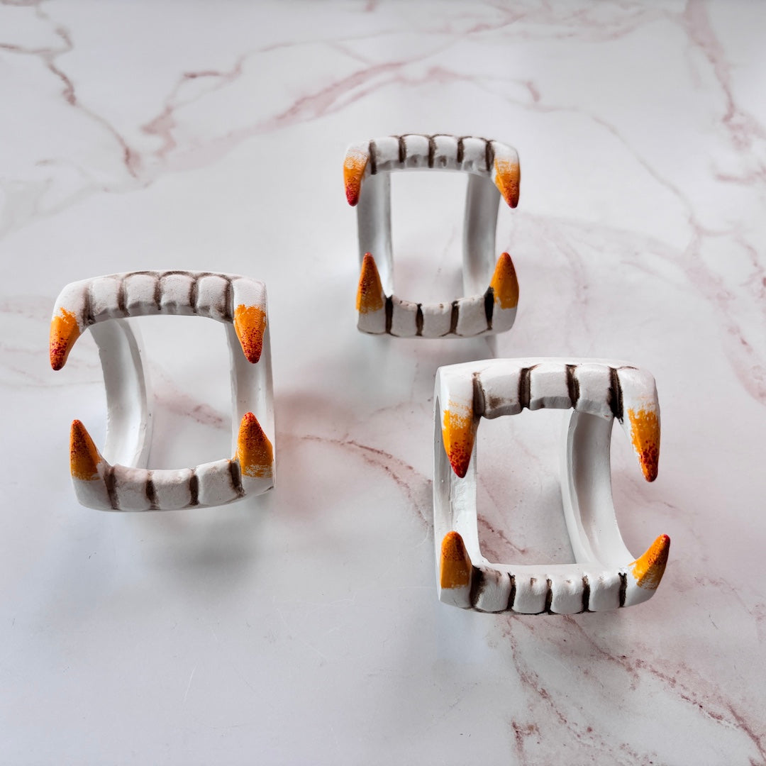 Three white ceramic vampire fang napkin rings arranged on a white marble surface with pink veining.