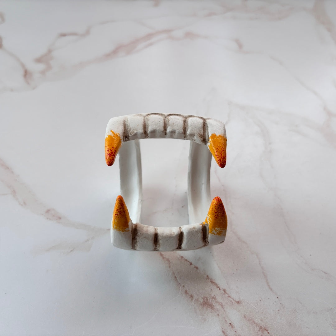A ceramic vampire fang napkin ring displayed on a white marble surface. The ring features white ceramic teeth with brown segmented details and vibrant orange-yellow tips.