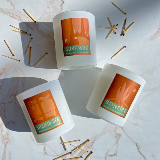 Three candles by Feather & Coco labeled 'Surf Wax', 'Vitamin Sea', and 'Bonfire' on a marble surface with matches.