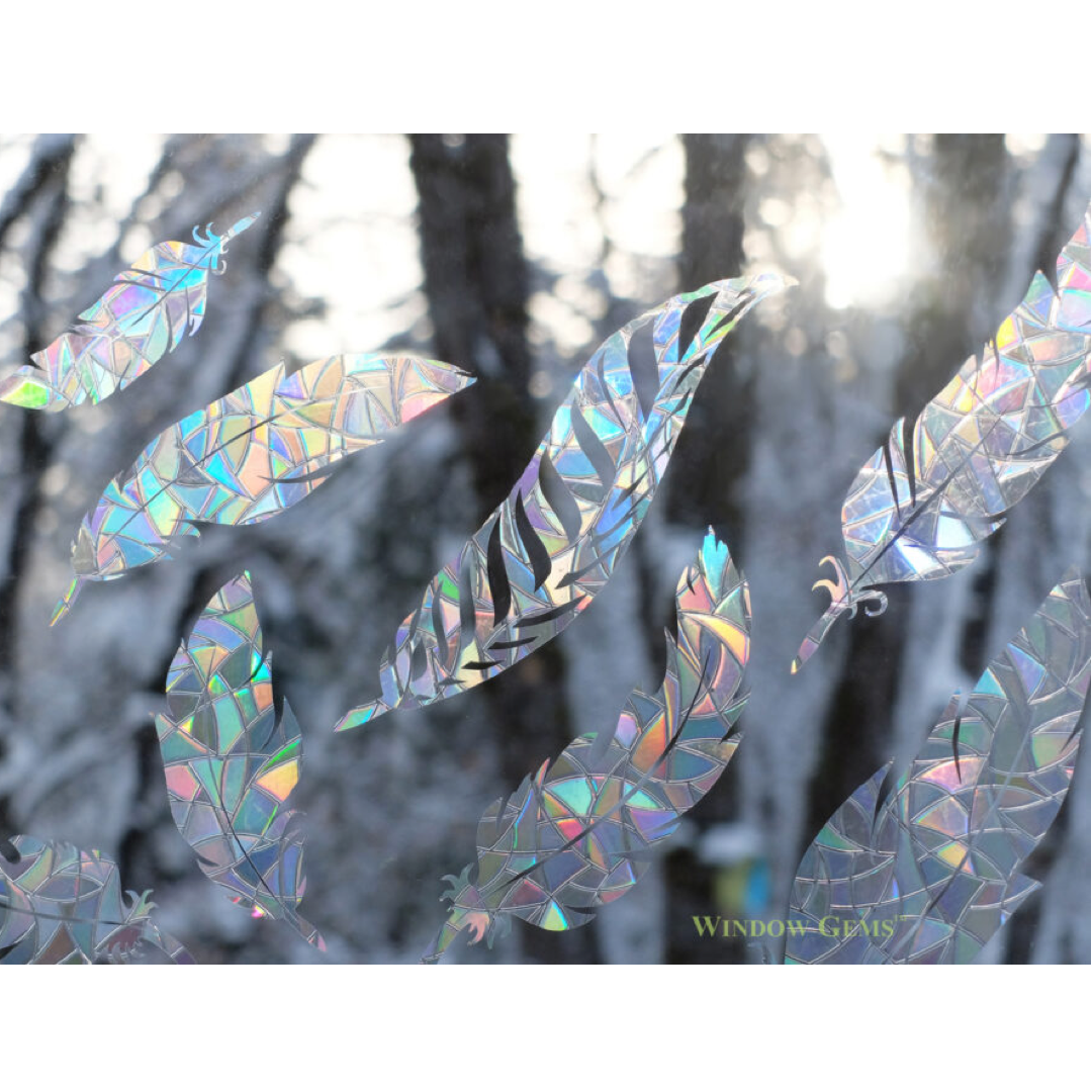 Iridescent holographic feather-shaped window clings displayed on a snowy forest window background, reflecting rainbow colors.