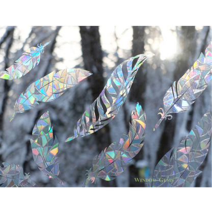 Iridescent holographic feather-shaped window clings displayed on a snowy forest window background, reflecting rainbow colors.