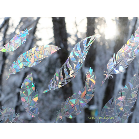 Iridescent holographic feather-shaped window clings displayed on a snowy forest window background, reflecting rainbow colors.