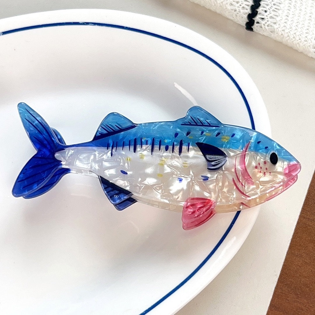 A fish-shaped hair clip with a transparent clear body, blue dorsal and fin areas, and pink lower details, resting on a white oval plate with a blue rim.