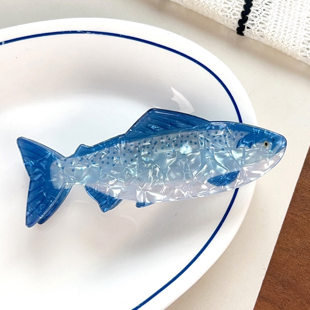 A translucent blue and clear fish-shaped decorative object resting on a white oval plate with a blue rim, placed on a light tabletop with a small wood surface visible at the corner.
