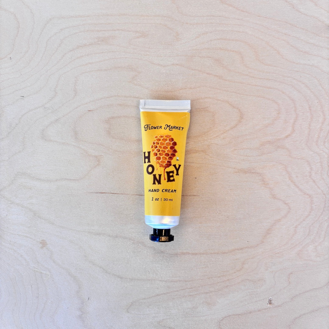 A squeeze tube of Flower Money Honey Hand Cream placed on a light wood grain surface.