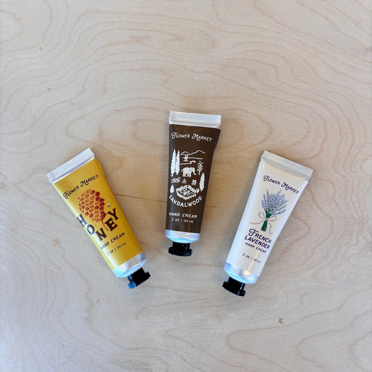 Three hand cream tubes from Flower Market are arranged on a light wood grain background. 