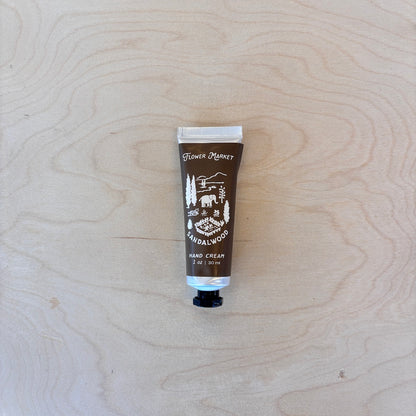 A squeeze tube of Flower Market Hand Cream in Sandalwood scent, placed vertically on a light-colored wood grain background.