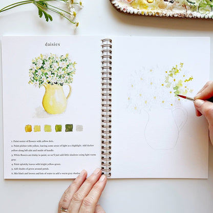 Open art book with daisies illustration and hand holding a paintbrush