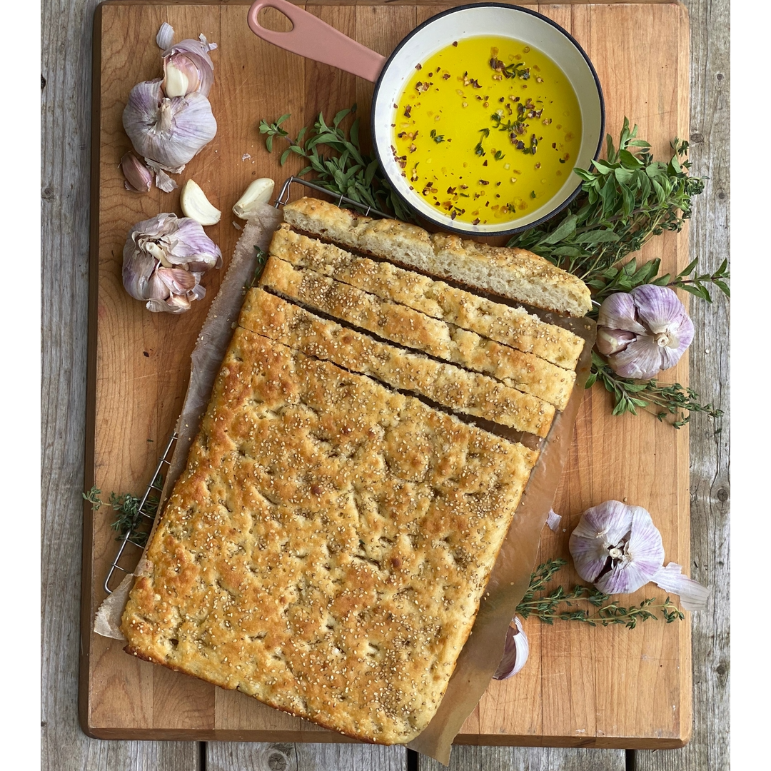 A rectangular loaf of focaccia bread on a wooden cutting board, sliced into thick strips. A small bowl filled with yellow olive oil and herbs sits at the top, surrounded by whole garlic bulbs, thyme sprigs, and a knife on a rustic wooden table.