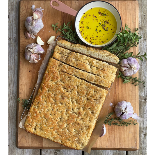 A rectangular loaf of focaccia bread on a wooden cutting board, sliced into thick strips. A small bowl filled with yellow olive oil and herbs sits at the top, surrounded by whole garlic bulbs, thyme sprigs, and a knife on a rustic wooden table.