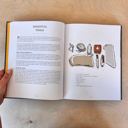 An open instructional book titled 'Essential Tools' being held by a hand, showing printed text and illustrated tool diagrams on two pages against a light wood background.