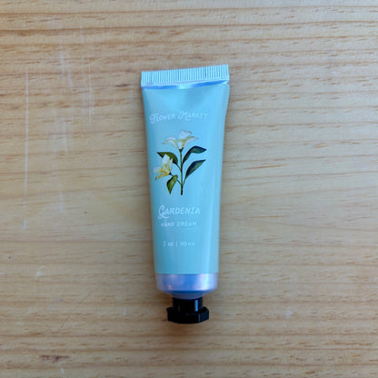 A light green squeeze tube of Flower Market Gardenia Hand Cream with a black cap, placed on a light wood grain surface.