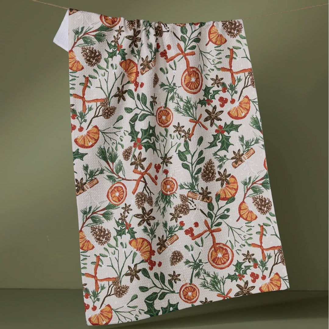 A white tea towel with a festive Christmas holly and citrus print hangs from a string against a muted olive green background.