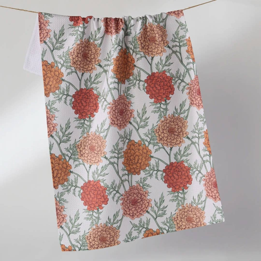 A white kitchen towel with a repeating pattern of orange, red, and peach marigold flowers and green leaves, hanging from a string against a light gray background.