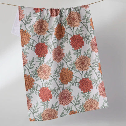 A white kitchen towel with a repeating pattern of orange, red, and peach marigold flowers and green leaves, hanging from a string against a light gray background.