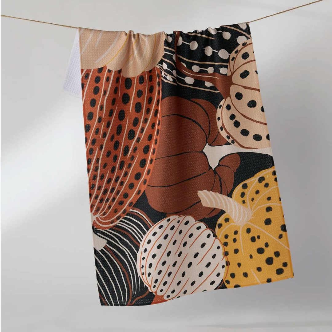 a tea towel hanging from a string on a white background. The fabric features a stylized autumn pumpkin pattern with multiple designs including solid orange, yellow, black-and-white polka dots, and brown-and-white stripes.