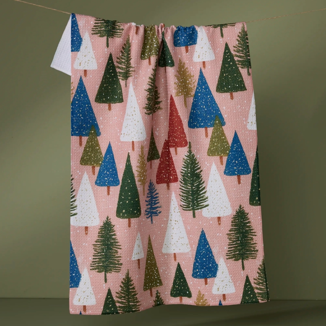 A pink kitchen towel with a repeating Christmas tree pattern in green, blue, white, and red, suspended against a plain olive-green background.