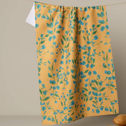 A mustard yellow kitchen towel with a blue and green floral pattern hanging from a string against a neutral beige background.