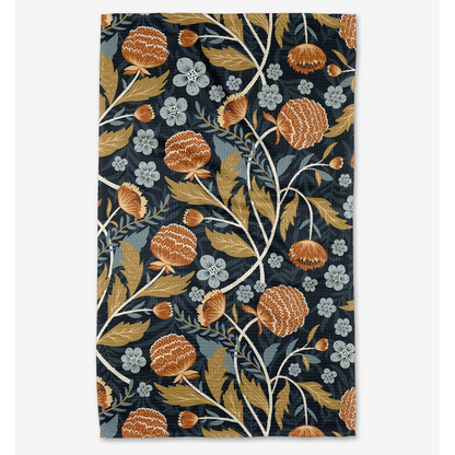 A tea towel with a dense botanical pattern, featuring orange-brown thistle-like flowers, muted gold leaves, and small pale blue blossoms on a dark navy background, shown flat against a light gray backdrop.