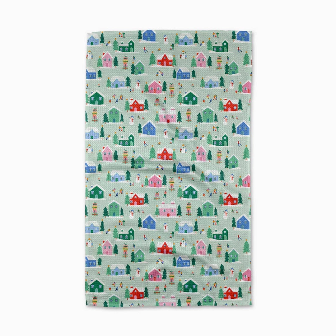 A tea towel with a repeating winter village pattern featuring colorful cabins, trees, and figures on a pale mint green background, shown flat against a white background.