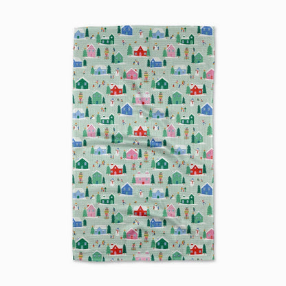 A tea towel with a repeating winter village pattern featuring colorful cabins, trees, and figures on a pale mint green background, shown flat against a white background.