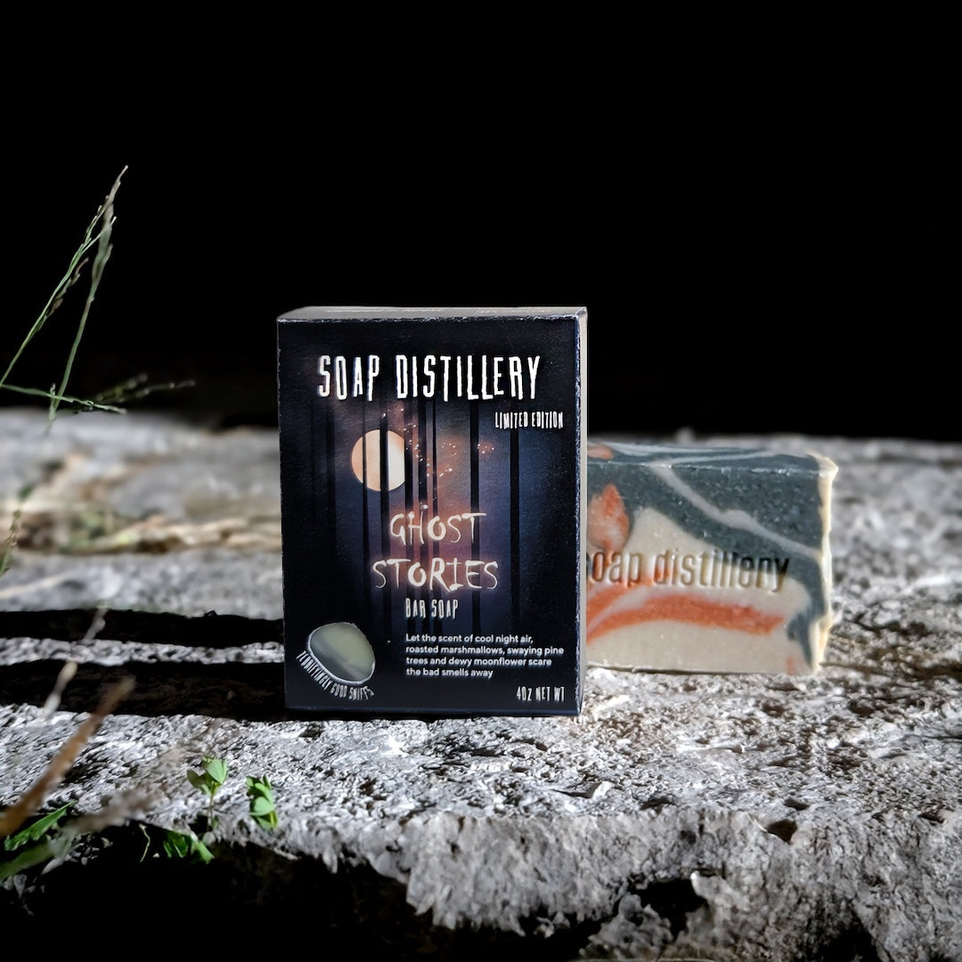 A boxed bar of soap labeled 'Soap Distillery Ghost Stories Limited Edition' stands upright on a rough stone surface, with a marbled bar of soap placed behind it against a dark background.